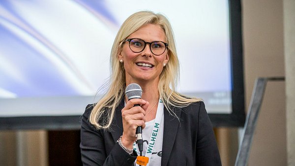 Blonde junge Frau mit Brille. Sie trägt Business-Kleidung und spricht in ein Mikrofon. Eine Präsentation zum Thema Fliegen und Assessment Tools.