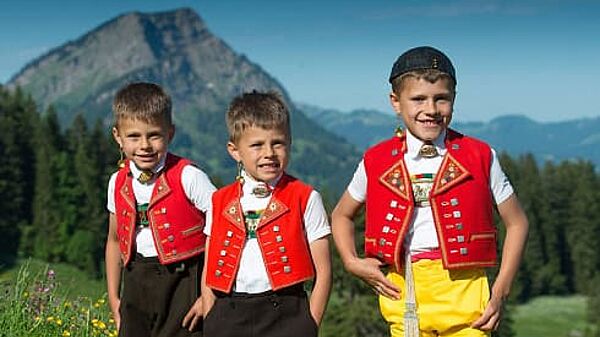 Drei Buben in Trachtenkleidung stehen auf der Alm. Sie blicken frühlich Richtung Kamera. Werbeaufnahme der Firma Appenzeller Alpenbitter AG.