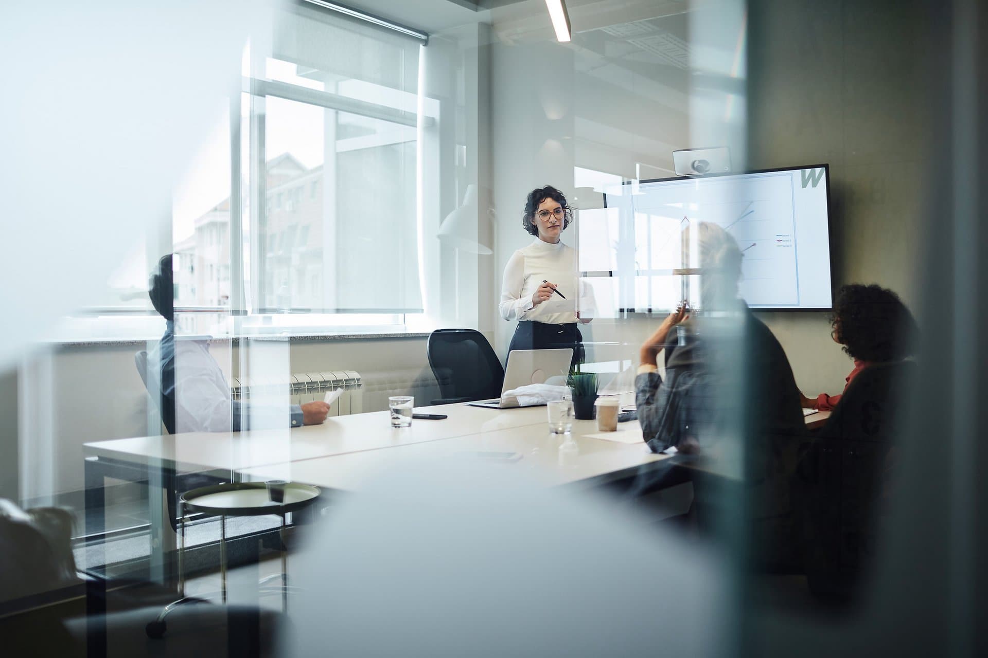 Wir sehen durch eine große Glasscheibe ins Büro. Eine junge Frau führt mit zwei Mitarbeiterinnen eine Management Consultancy durch.